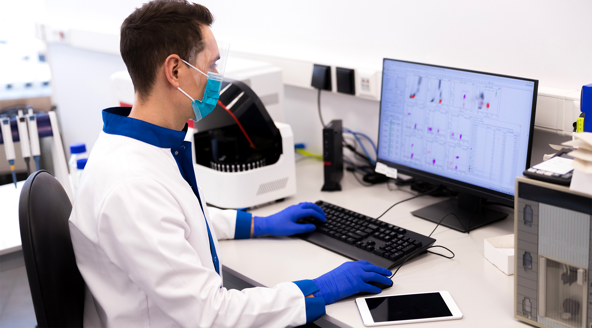 A medical researcher wears a mask and gloves while logging data on a desktop computer.