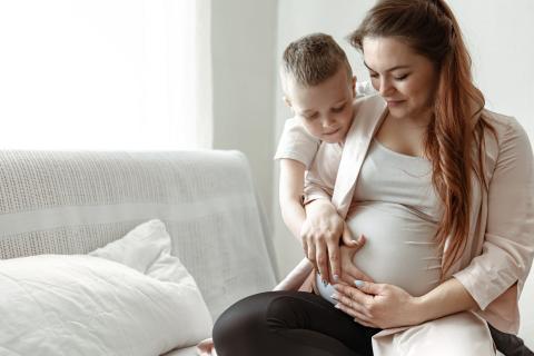 A child sits with a pregnant person. They both hold their hands to the pregnant person's stomach.